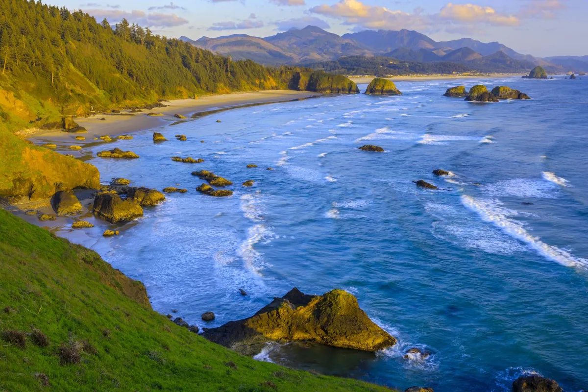 This image captures a tranquil coastal view with ocean waves gently rolling onto a sandy beach, framed by striking rock formations, lush trees, and distant mountains. The golden-hour lighting evokes the quiet beauty of sunset.