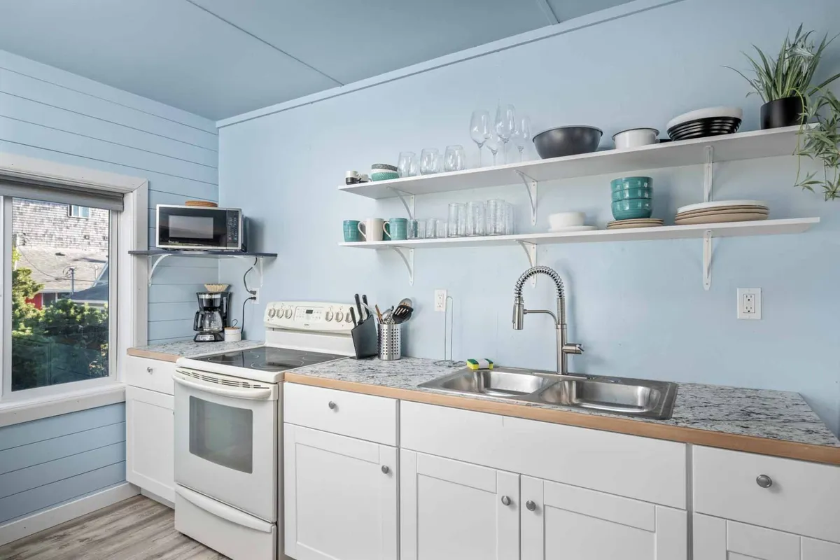 This image presents a bright kitchenette with a granite countertop, a kitchen sink, and shelves neatly arranged with dishware. The window brings in cheerful light, illuminating a space ideal for preparing simple meals, making coffee, or enjoying breakfast.