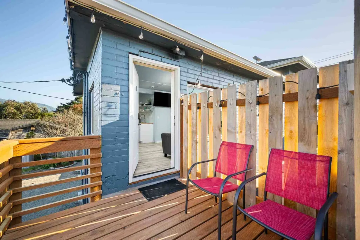 A cheerful exterior view of a wooden deck with bright red chairs and an open door, indicating easy access between indoor and outdoor areas. The space feels private yet sociable, perfect for relaxing, reading, or letting a pet lounge by your side.