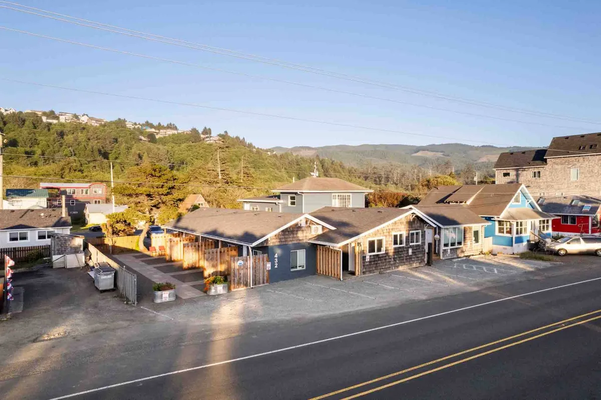 A row of colorful coastal houses surrounded by greenery, with a parking area in the foreground and a mountain range providing a scenic backdrop. The relaxed setting evokes the charm of small coastal communities and the sense of discovery while exploring local neighborhoods.