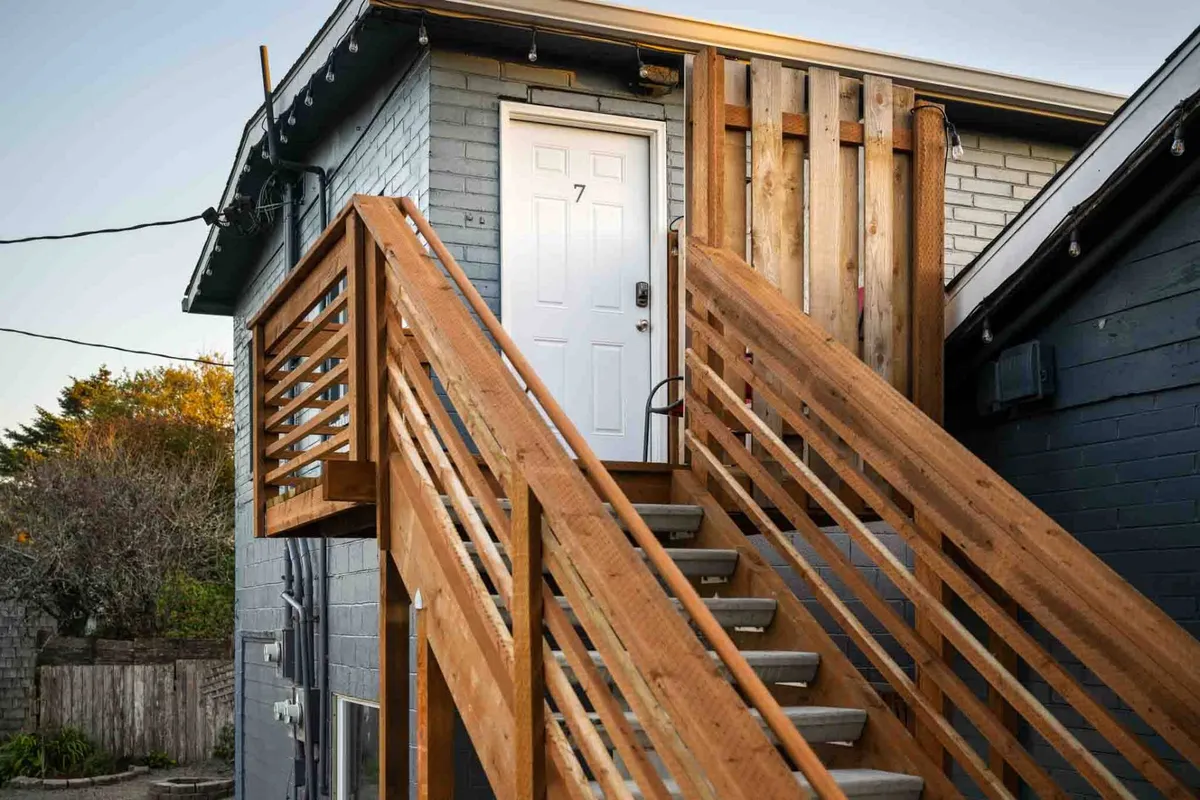 The image captures the welcoming entrance to one of the units, featuring a wooden staircase, abundant greenery, and a numbered door. The outdoor setting sets a serene, homey mood and suggests a private space to relax after a beach adventure.