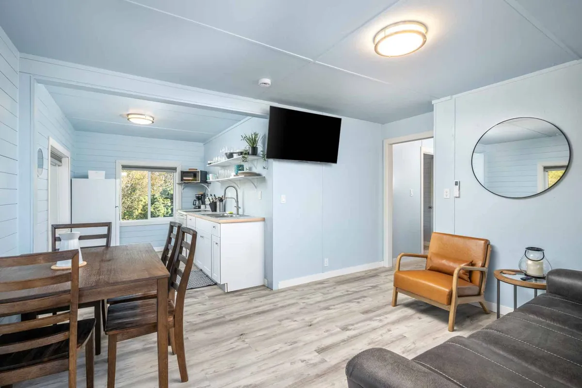 A welcoming, open-plan space featuring a dining table, kitchen area, a comfortable sofa, wall-mounted TV, and a round mirror. The area is bright, with light streaming in, and all core living functions are within view—dining, relaxing, and socializing.