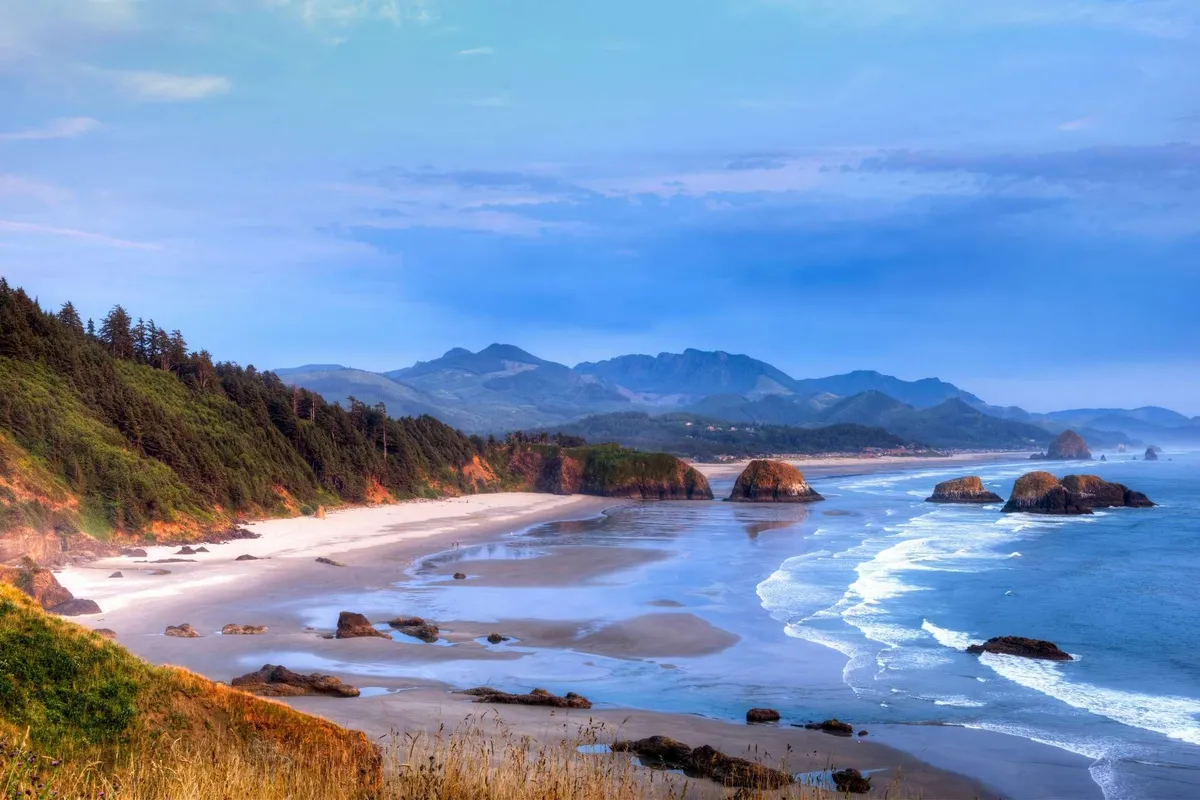 This wide-angle view showcases the stunning Rockaway coastline, with sandy stretches, ocean waves, dramatic rock formations, and mountains in the distance. The scene is serene and expansive, capturing the natural beauty and adventure that awaits just beyond the cottages.
